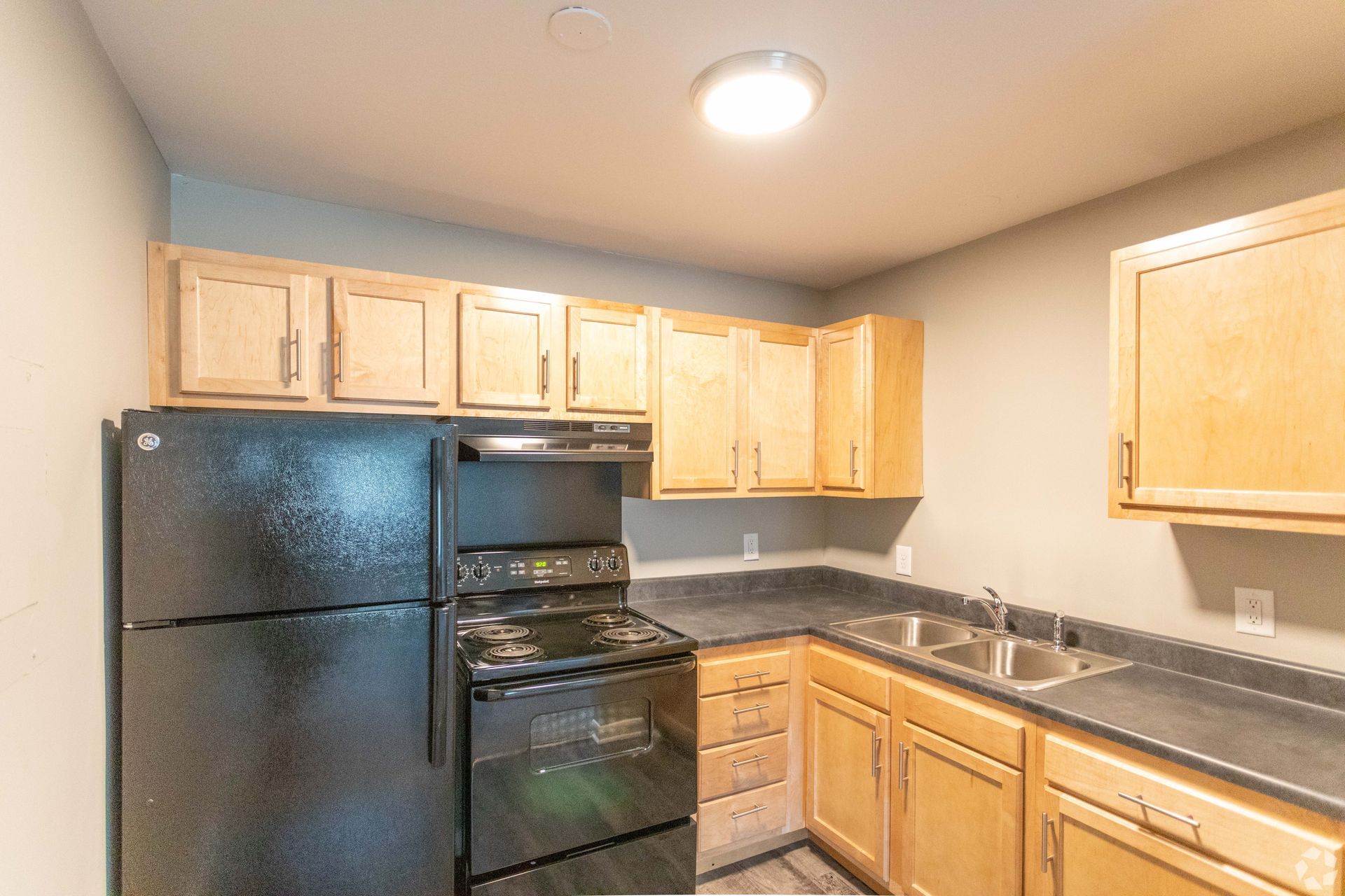 A kitchen with light wood cabinets, black appliances, a dark countertop, and a double basin sink.