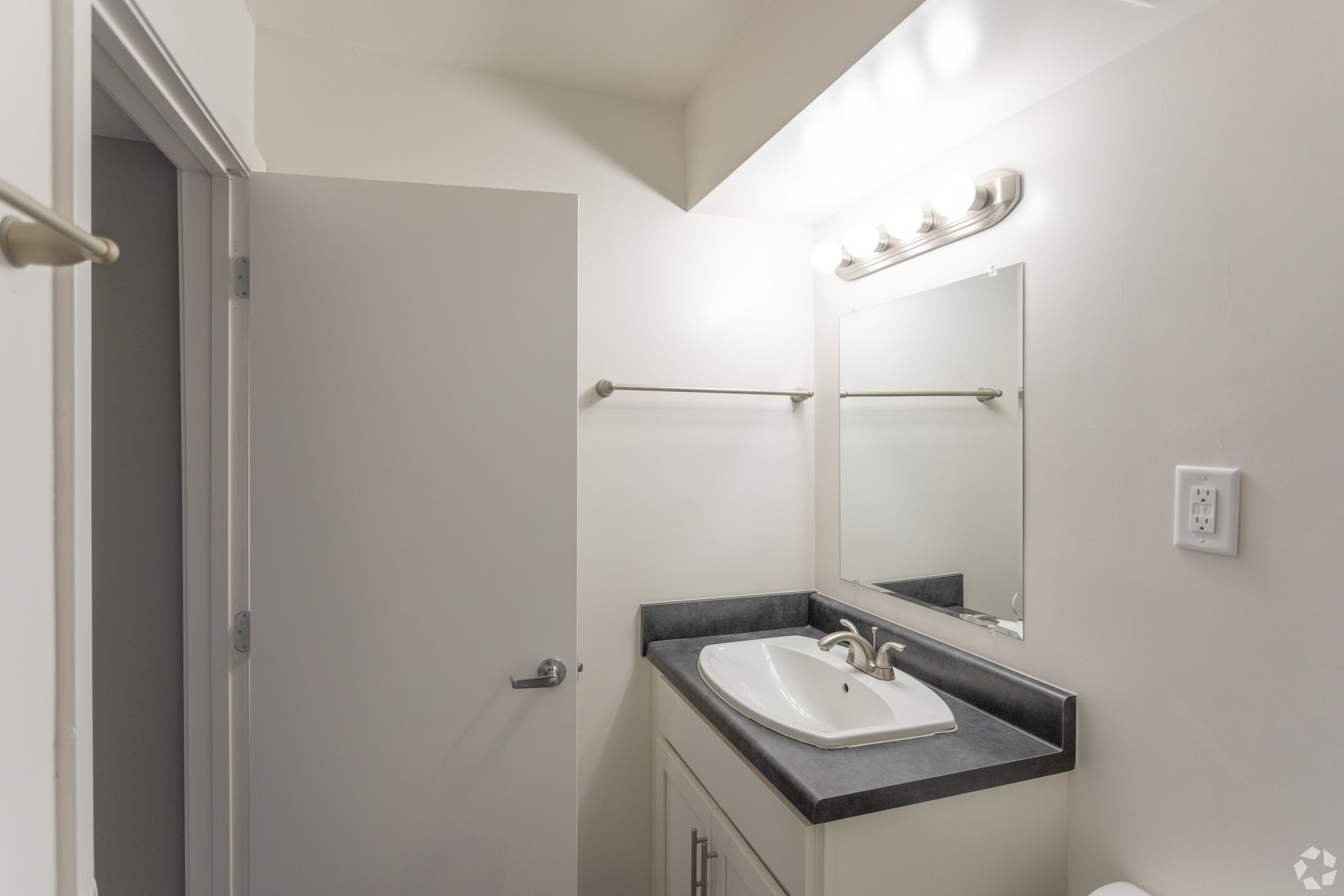 A neutral bathroom featuring a white vanity with a dark gray countertop, a sink, a mirror, and a wall-mounted light fixture.