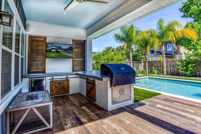 outdoor kitchen by pool with palm trees lining yard. stainless grill and peninsula, wooden shutters and white lower storage