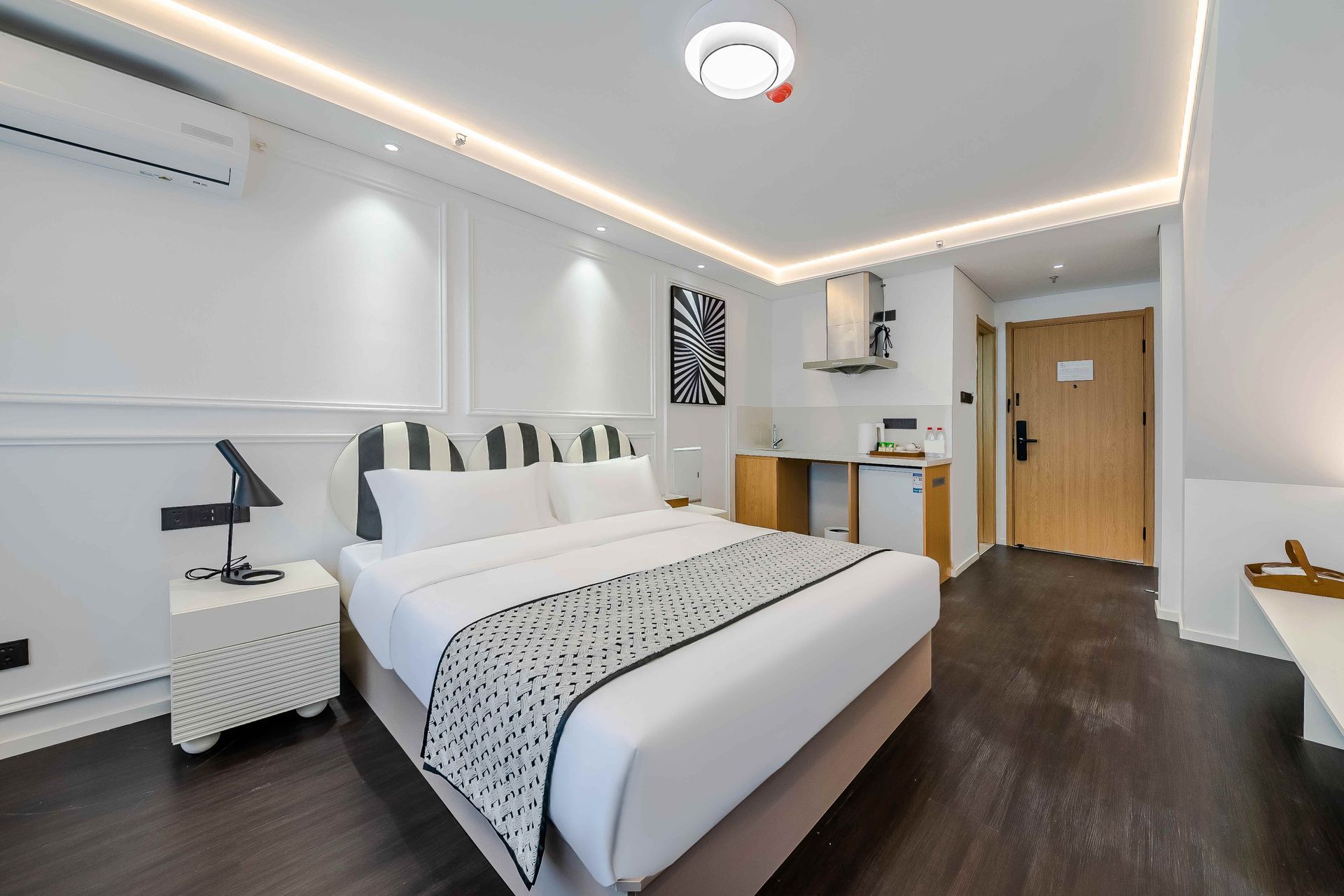 Modern bedroom with white bedding, dark wood floors, desk, and kitchenette area
