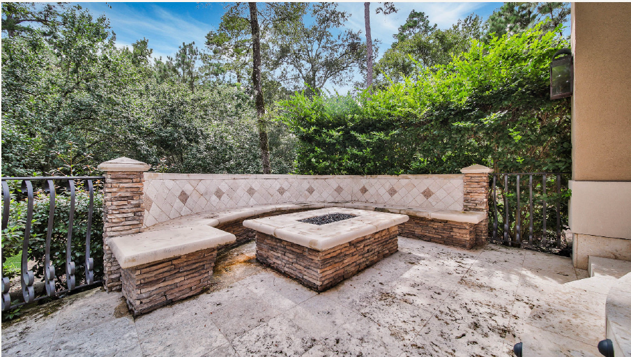 Stone patio with built-in bench and fire pit, surrounded by lush green trees and black iron railing.