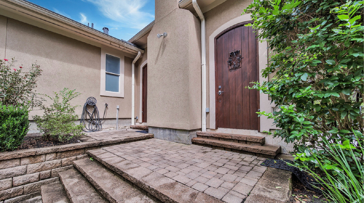 Front entrance of a beige house with a wooden door, stone steps, and landscaped shrubs.