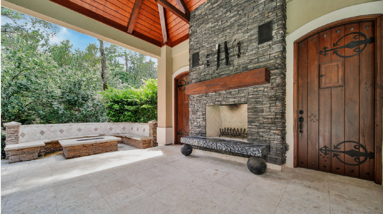 Covered stone patio with fireplace, wood door, bench seating, and wooded view beyond the railing