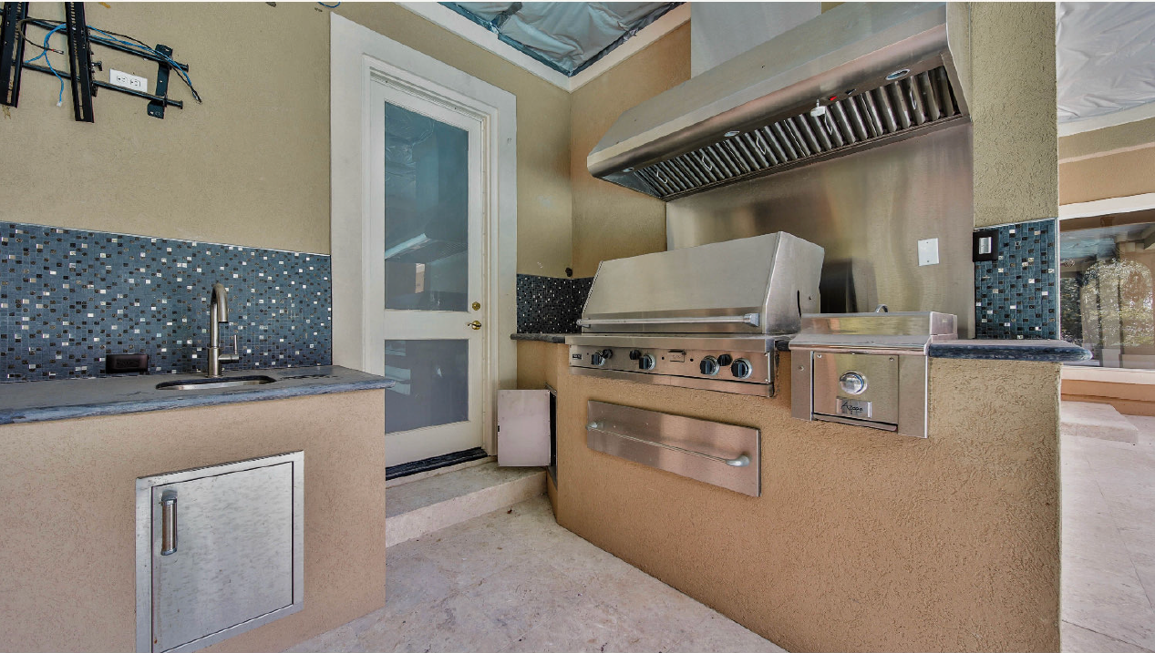 Modern outdoor kitchen with built-in grill, stainless vent hood, tan counters, and a glass door at left