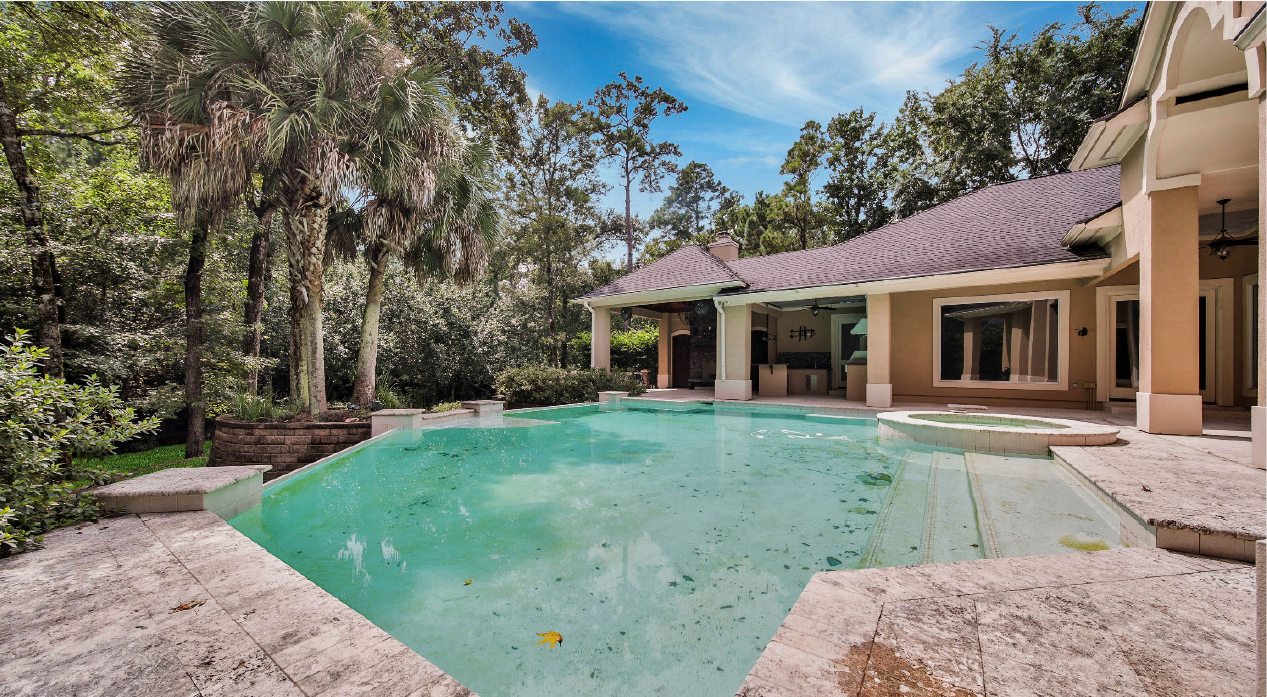 Backyard pool with turquoise water beside a beige house and tall trees