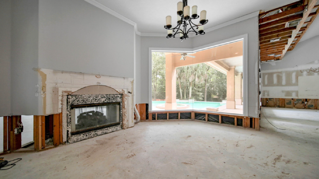 Spacious unfinished living room with fireplace, chandelier, and large bay window overlooking a backyard.
