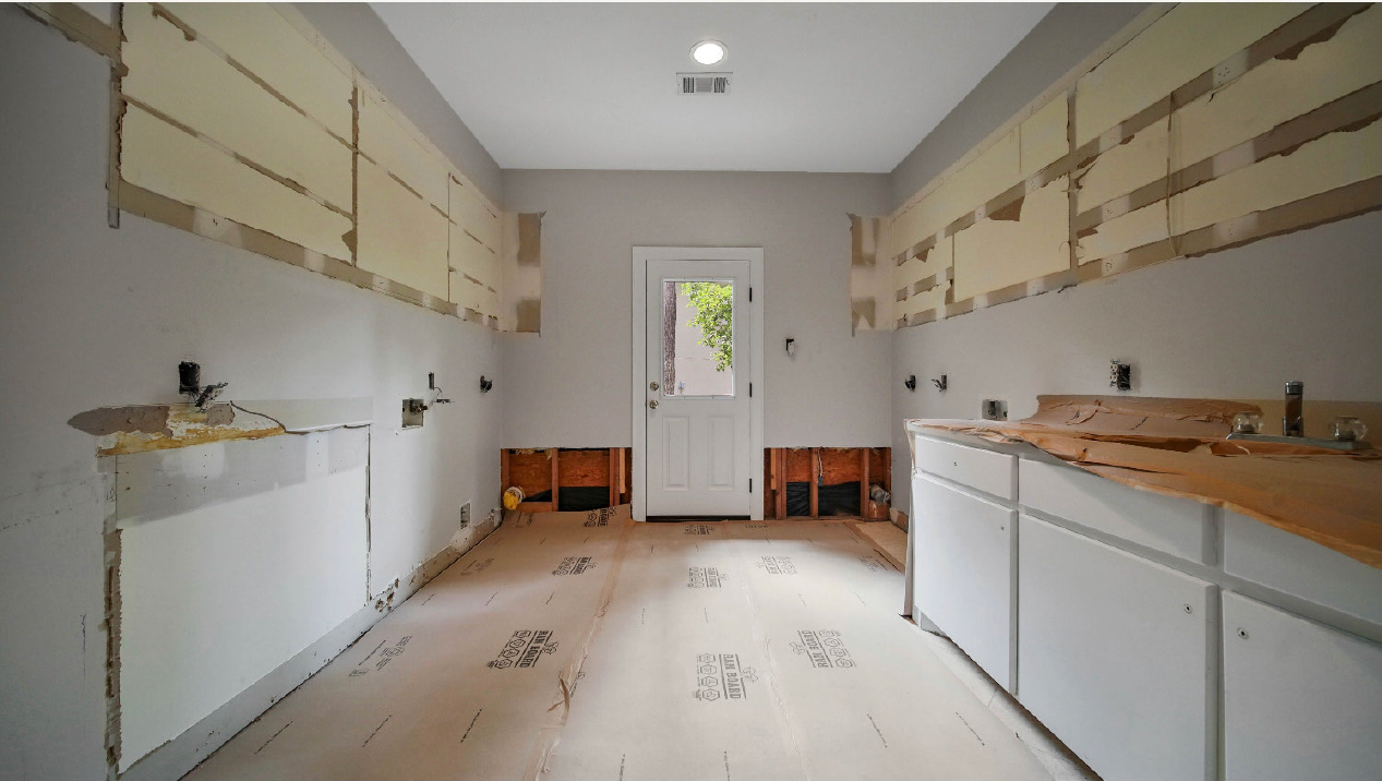 Empty kitchen under renovation with white cabinets, exposed wall patches, and a door at the back.