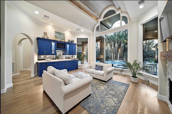 Bright modern living room with blue kitchen, cream sofas, arched windows, and forest view