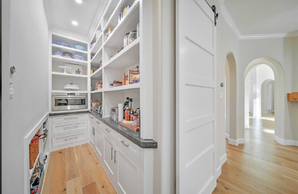 Bright white pantry with open shelves, cabinets, and wood floors in a hallway.
