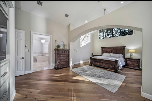 Spacious bedroom with vaulted ceiling, arched window, wood floor, king bed, dresser, and area rug.