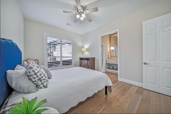 Bright bedroom with a bed, ceiling fan, window shutters, and an open doorway to a bathroom.