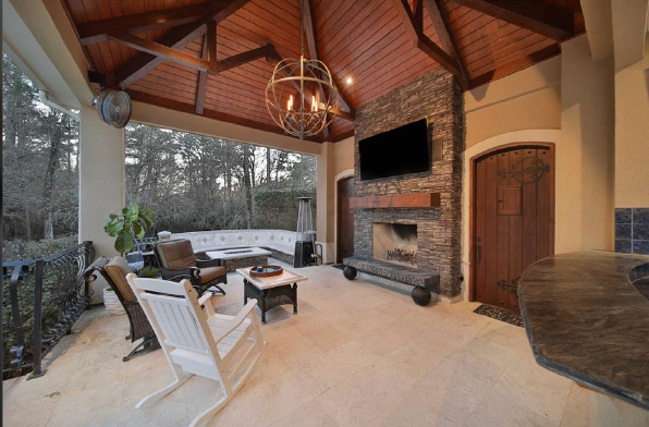 Covered outdoor patio with wood ceiling, stone fireplace, TV, and white chairs overlooking a wooded yard