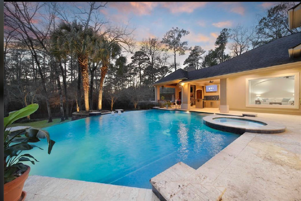 Backyard pool beside a lit modern home at dusk, with palm trees and a glowing blue water surface