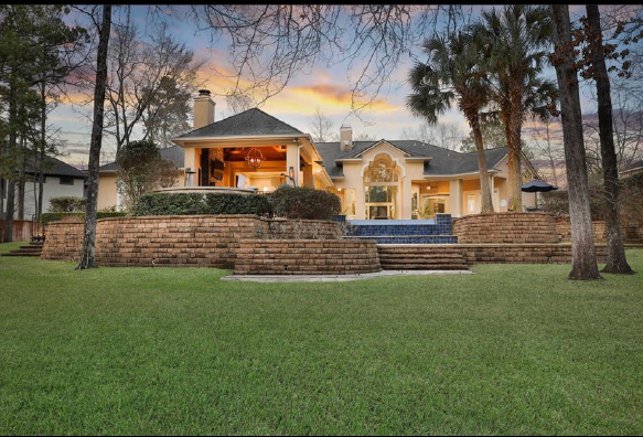 Large tan house with lit porch at dusk, behind a stone retaining wall and grassy lawn.