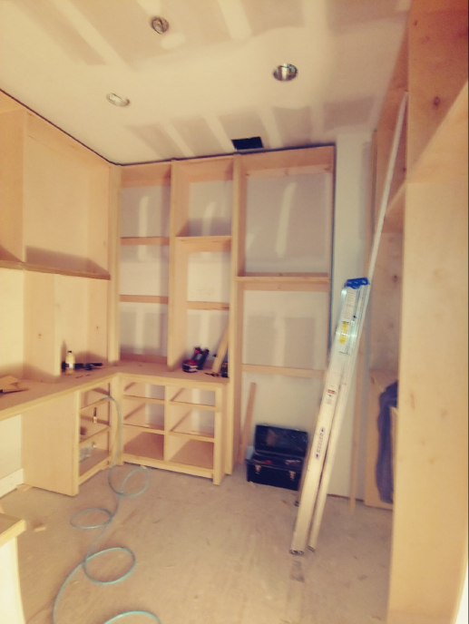 Partially built kitchen cabinets and shelving in an unfinished room with ladder and tools.