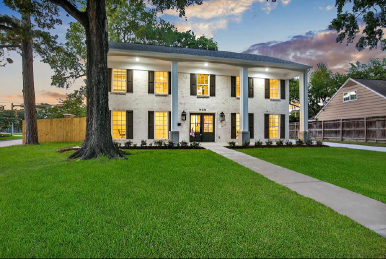 Two-story white colonial-style house with lit windows, large lawn, and a driveway at dusk