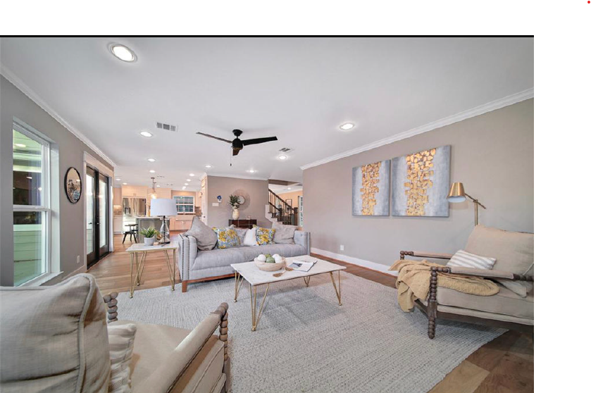 Bright living room with gray sofa, white coffee table, wood accents, ceiling fan, and open kitchen in back