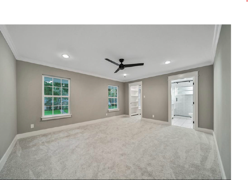 Empty beige bedroom with carpet, ceiling fan, windows, and an open door to a bathroom