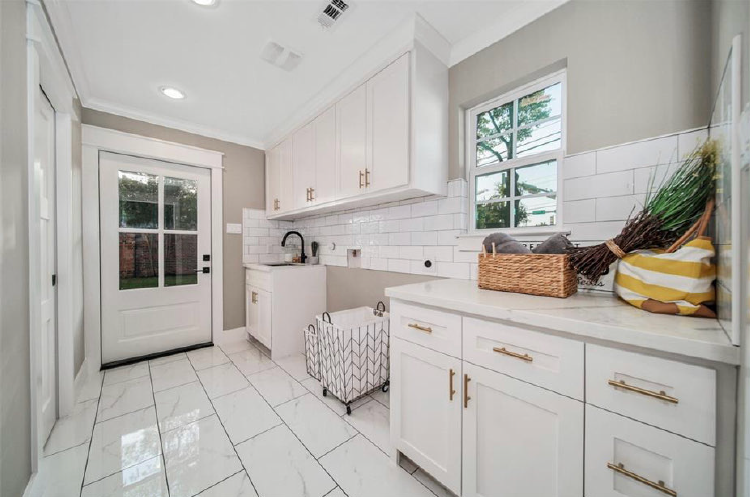 Bright laundry room with white cabinets, tiled floor, utility sink, and wicker baskets near a window