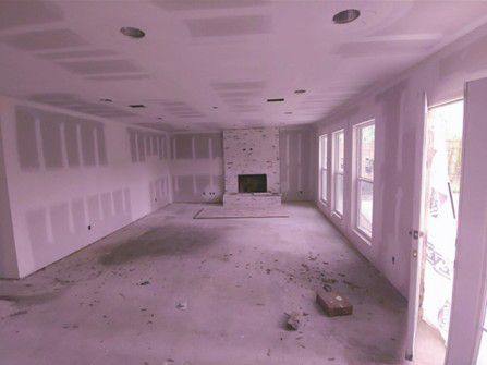 Empty unfinished living room with drywall patches, dusty floor, and large windows along one wall. Empty unfinished living room with drywall patches, dusty floor, and large windows along one wall.