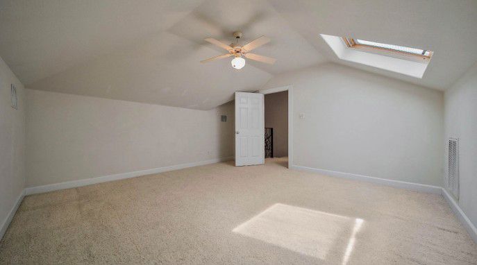 Empty carpeted attic bedroom with white walls, ceiling fan, skylight, and open closet door.