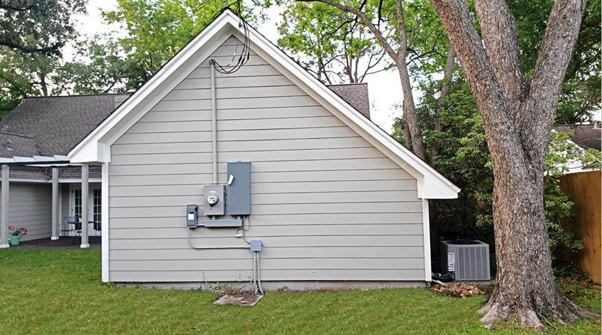 Side view of a gray house with utility meters, air conditioner, and a large tree in the yard