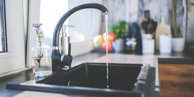 Black kitchen sink with running faucet and small vase of flowers by a bright window