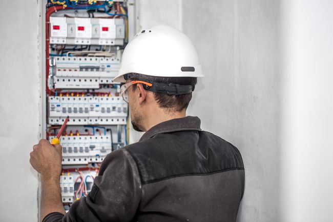 Electrician in a white hard hat working on an open electrical panel with wiring and circuit breakers.