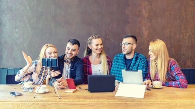 Five coworkers gathered around a laptop at a wooden table, smiling and chatting in an office setting