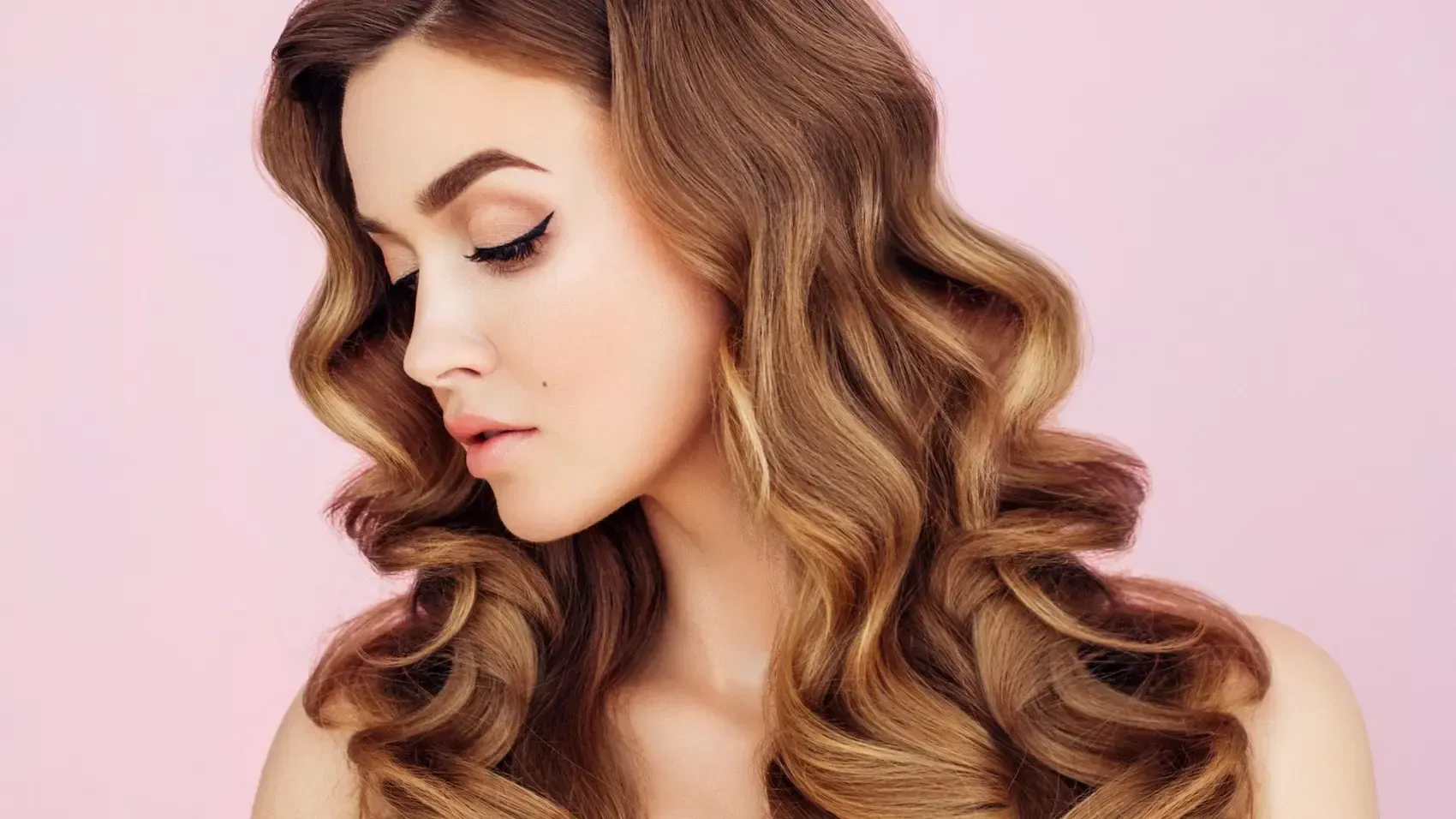 A woman with long curly hair is looking down on a pink background.