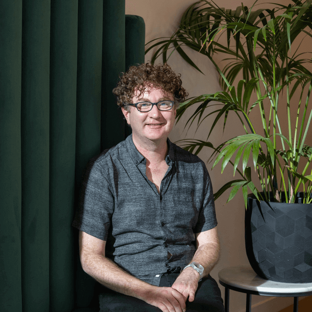 A man wearing glasses is sitting in front of a potted plant