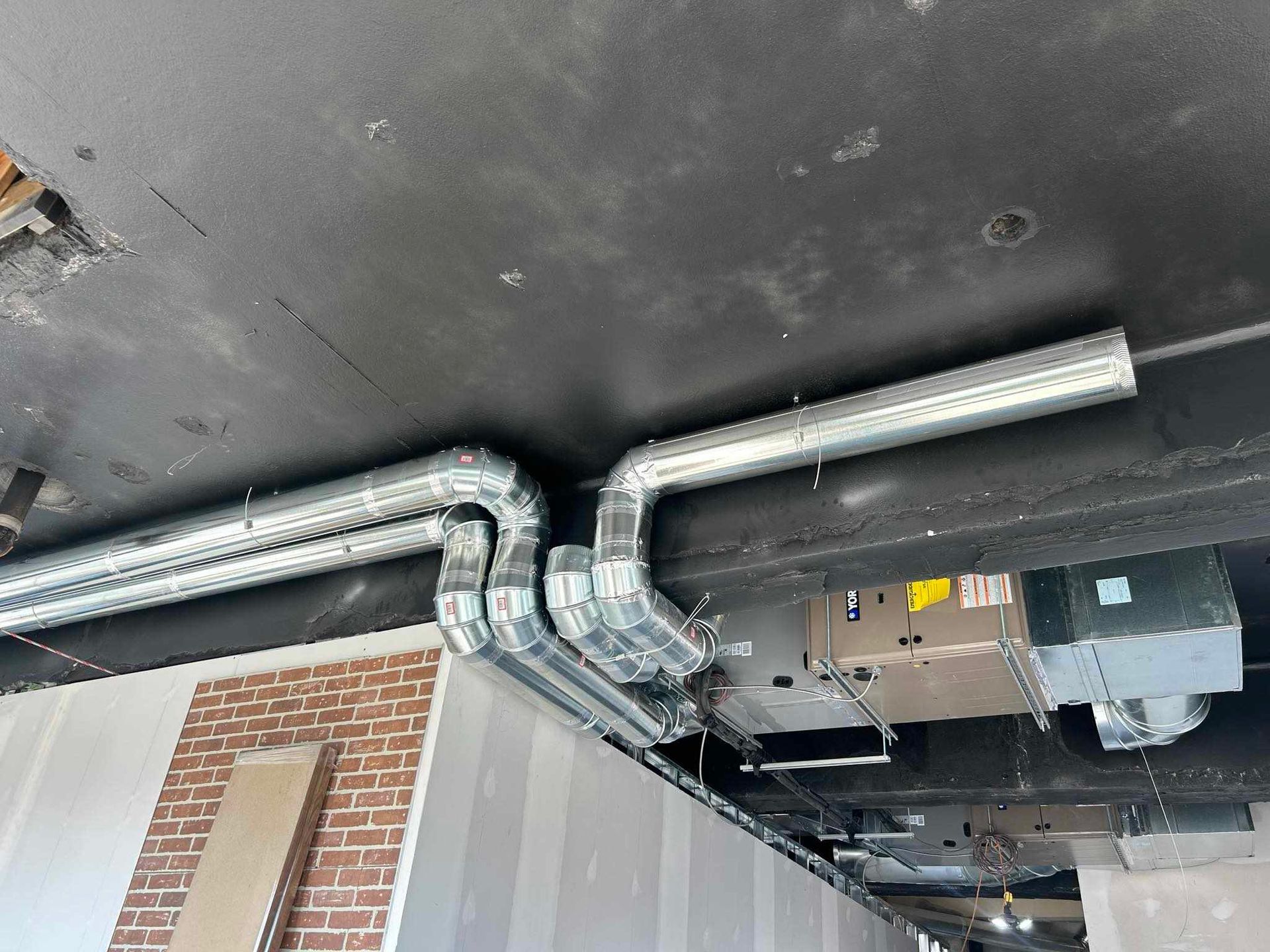 Conduits métalliques galvanisés installés au plafond, avec un mur de briques et d'autres matériaux de construction visibles.