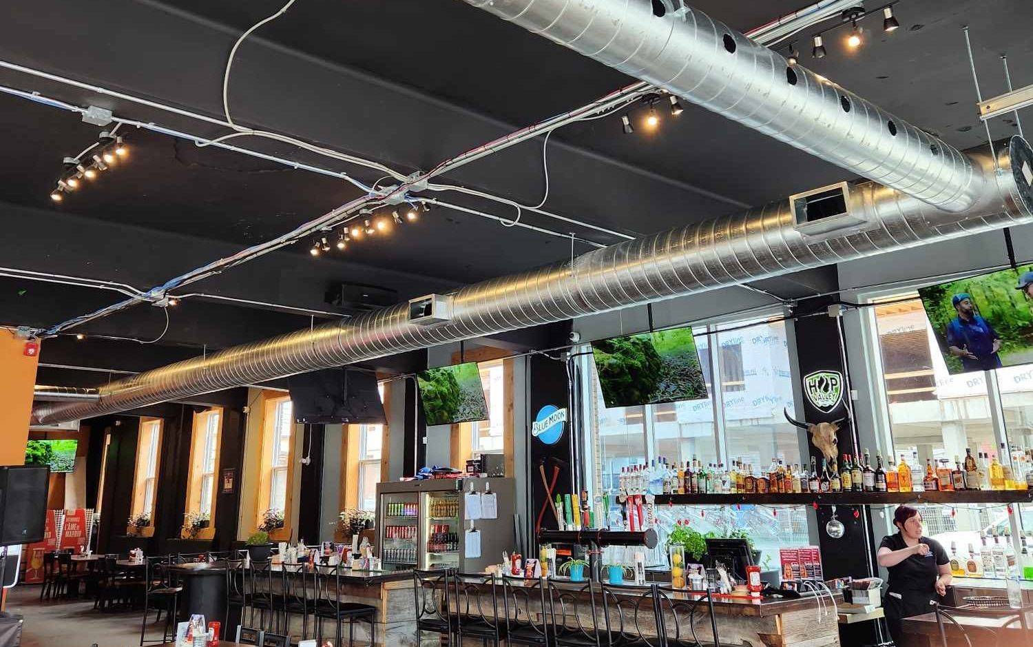 Intérieur d'un bar avec plafond noir, conduits d'aération apparents et téléviseurs. Un barman travaille derrière le comptoir.