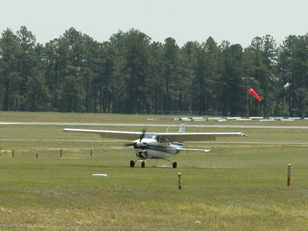Small white airplane on a grassy airfield with a red windsock and trees in the background