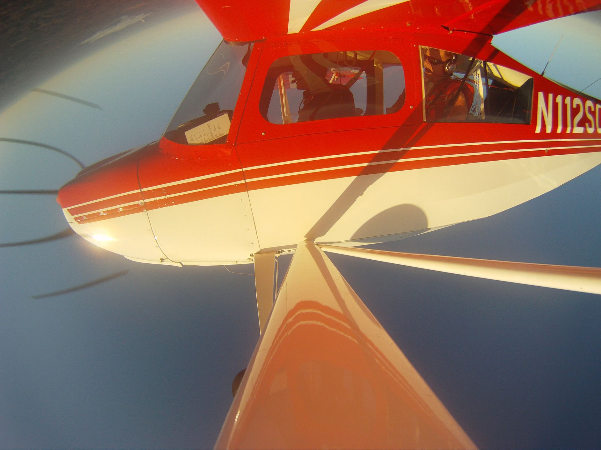 A view from the side of a red and white small airplane in flight, showing its wing, propeller, and registration N112SC.