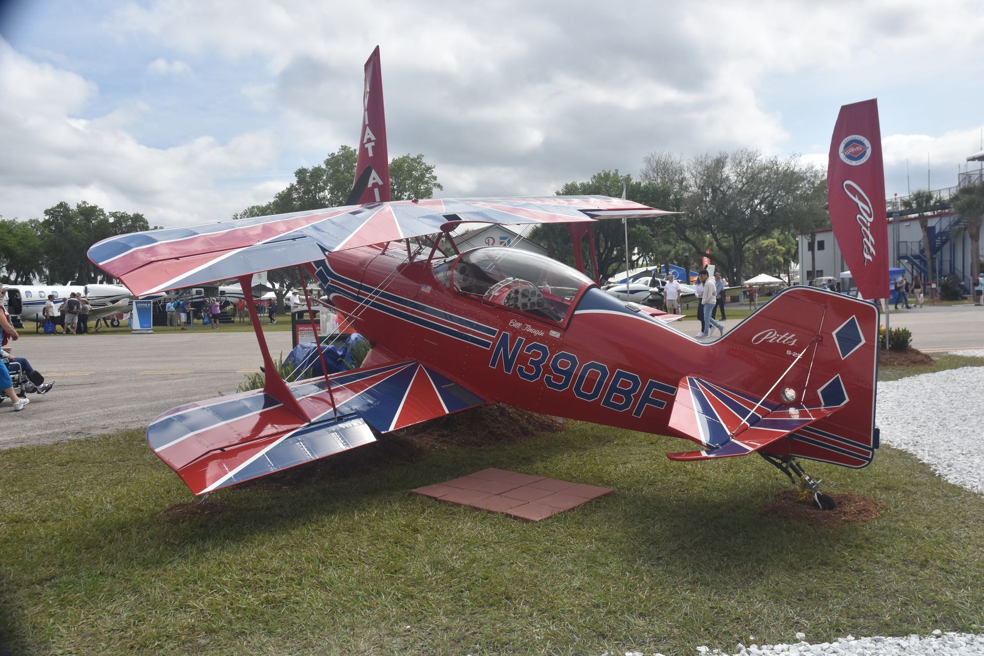 Red aerobatic biplane on grass display at an outdoor airshow, with spectators and flags nearby