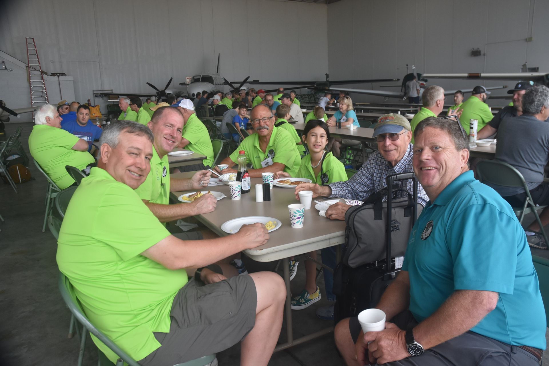 Group of people in bright green shirts seated at long tables in a warehouse-like hall, smiling and eating lunch