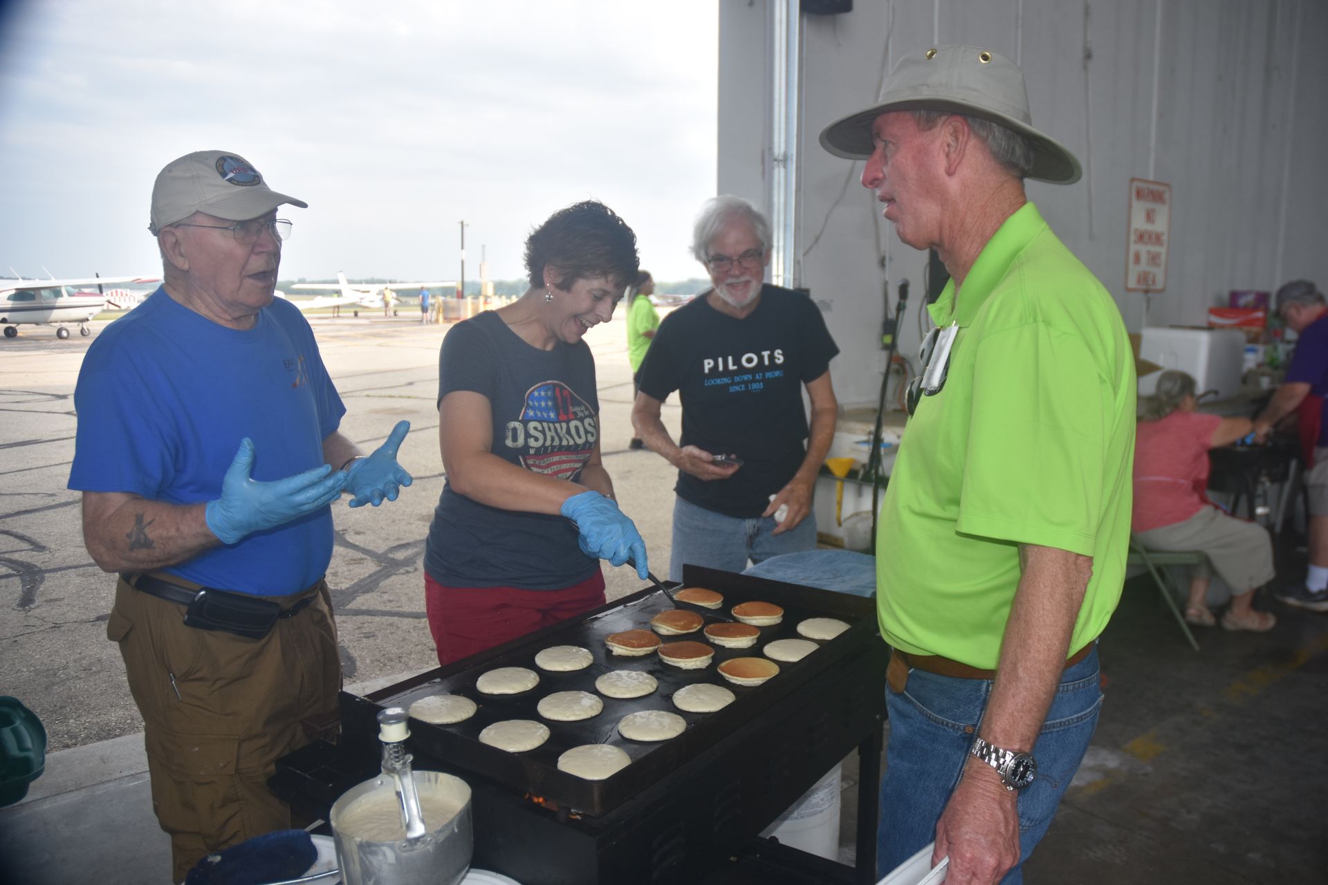 Four people in a hangar converse while preparing pancakes on a large griddle at an airport.