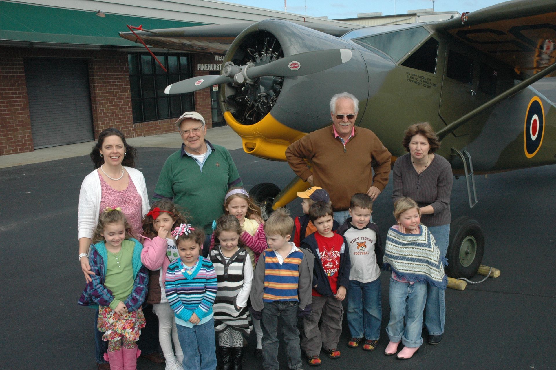 Group posing in front of a vintage airplane, with children lined up and adults standing behind them.