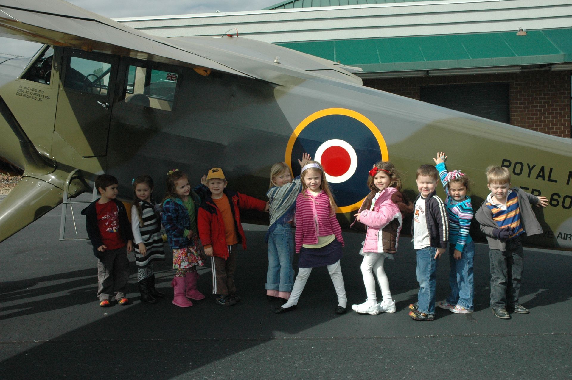 A line of ten young children posing and smiling in front of a Royal Air Force airplane on an outdoor runway.