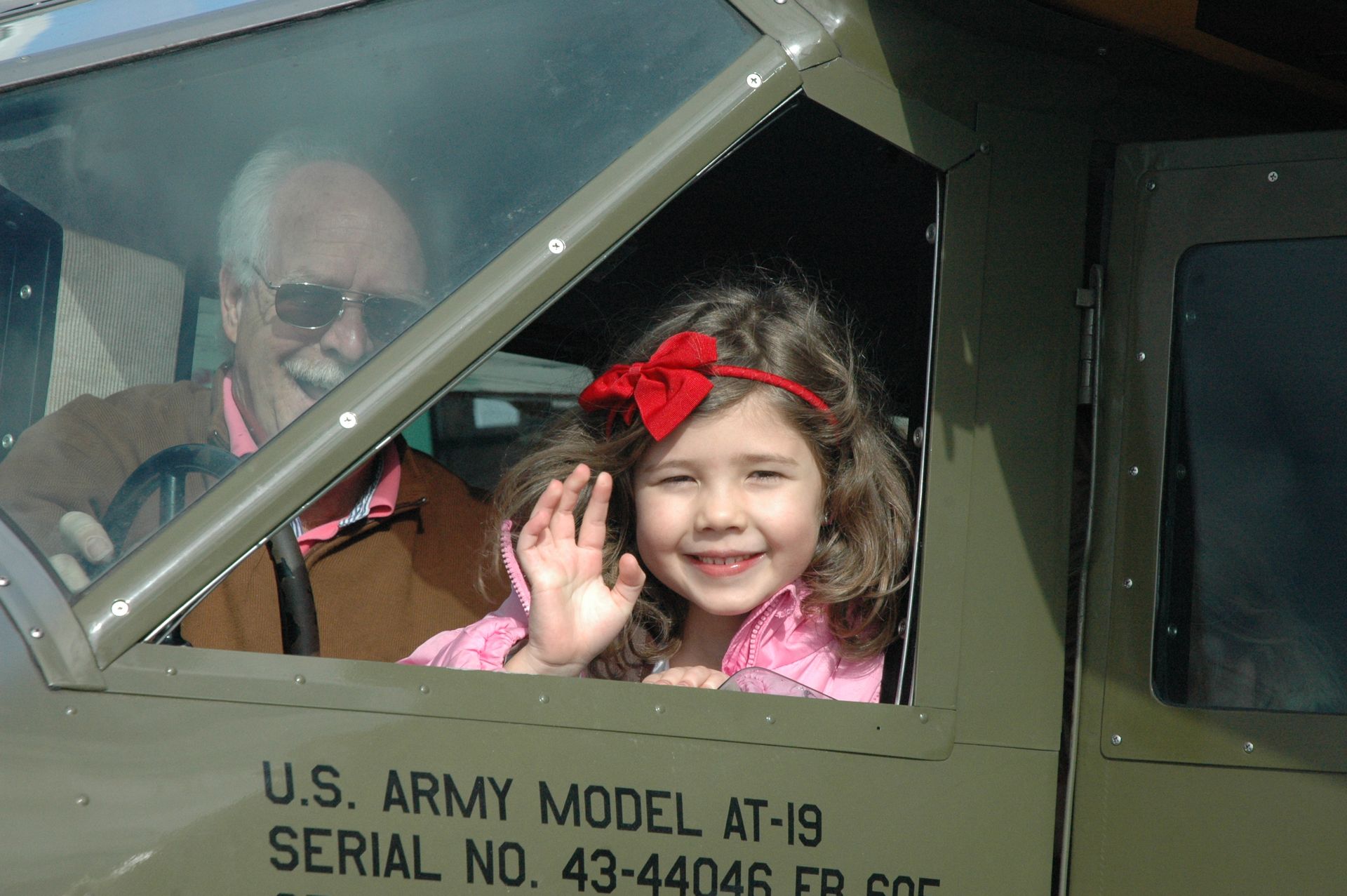 A young child with a red bow waves from inside the cockpit of a vintage olive green U.S. Army model AT-19 aircraft.