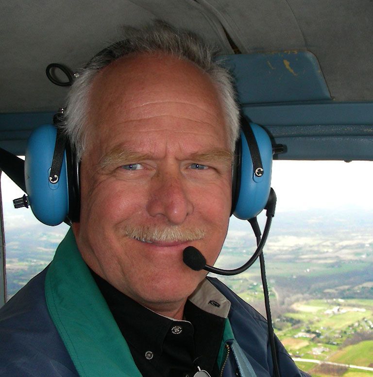 A person wearing a headset in the cockpit of a small aircraft, smiling with a rural landscape visible through the window.