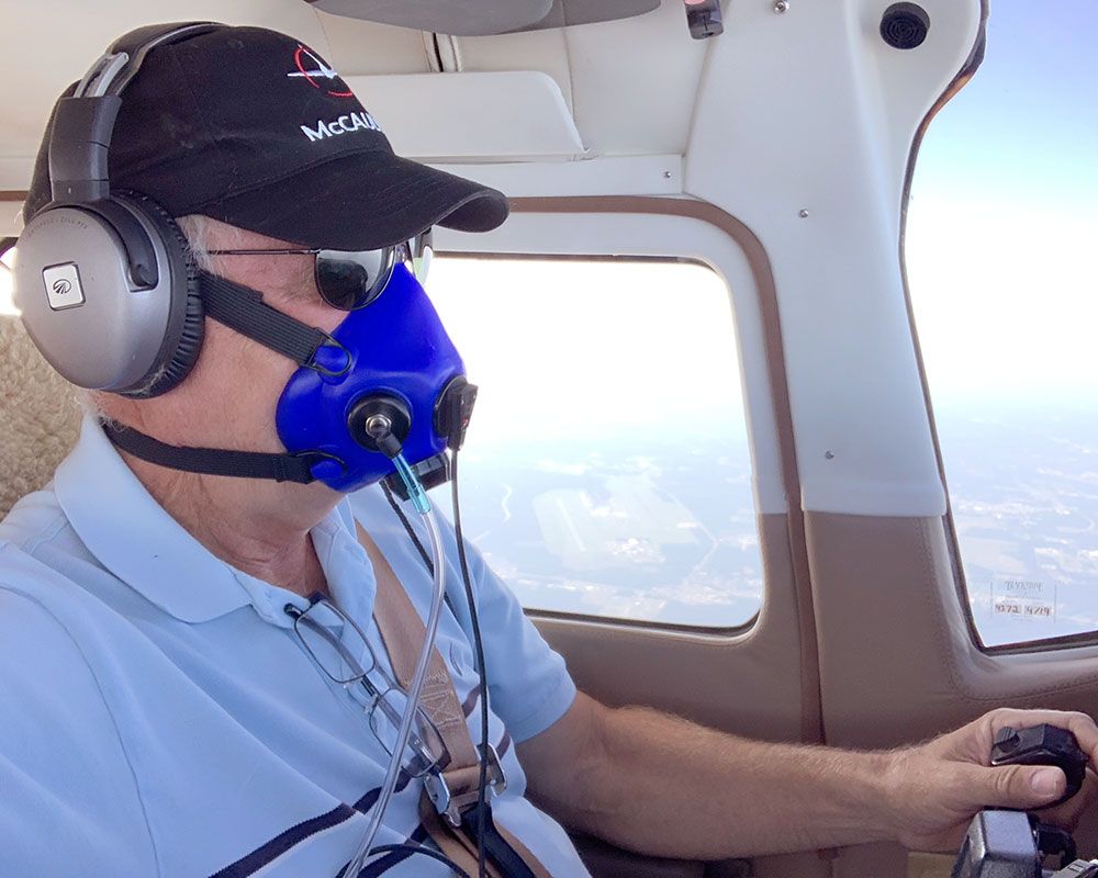 Pilot wearing blue oxygen mask in a small plane cockpit, looking out the window and holding the controls.