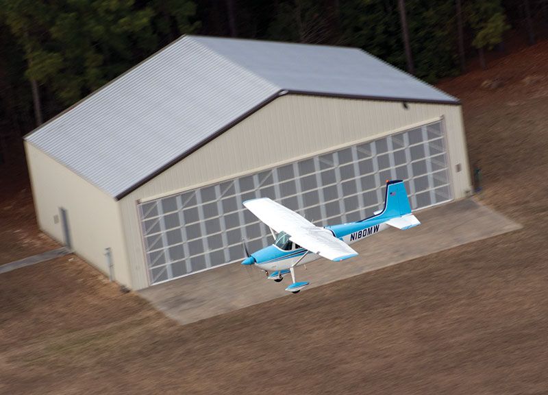 A small white and blue single-engine airplane flying in front of an aircraft hangar.