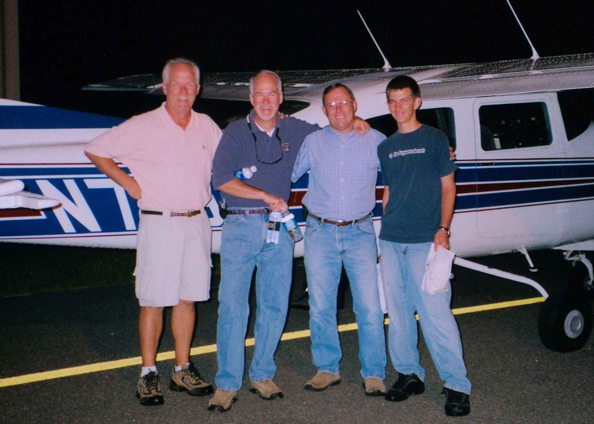 Four men stand smiling beside a small airplane at night.