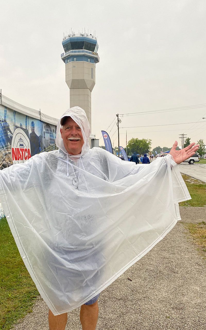 A smiling person wearing a clear plastic poncho with arms outstretched, standing outside near an airport control tower.