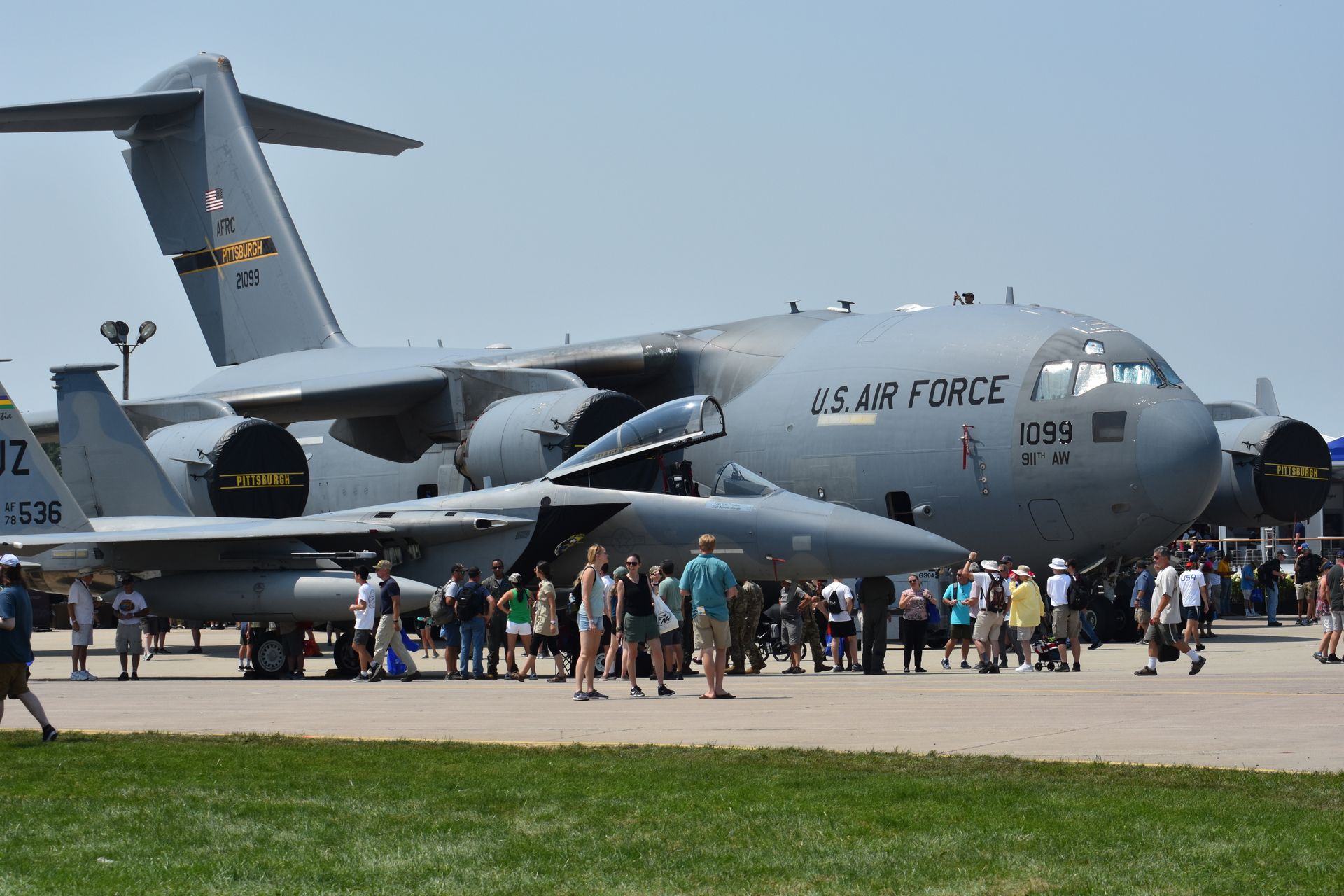 A crowd gathers on an airfield in front of a gray U.S. Air Force C-17 transport plane and an F-15 fighter jet.