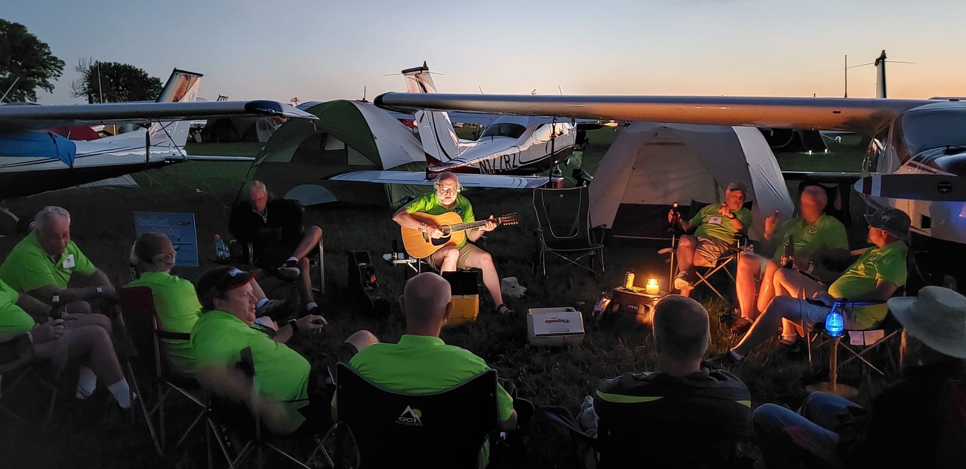 Campers seated around a glowing campfire at dusk beneath tents and parked vehicles.