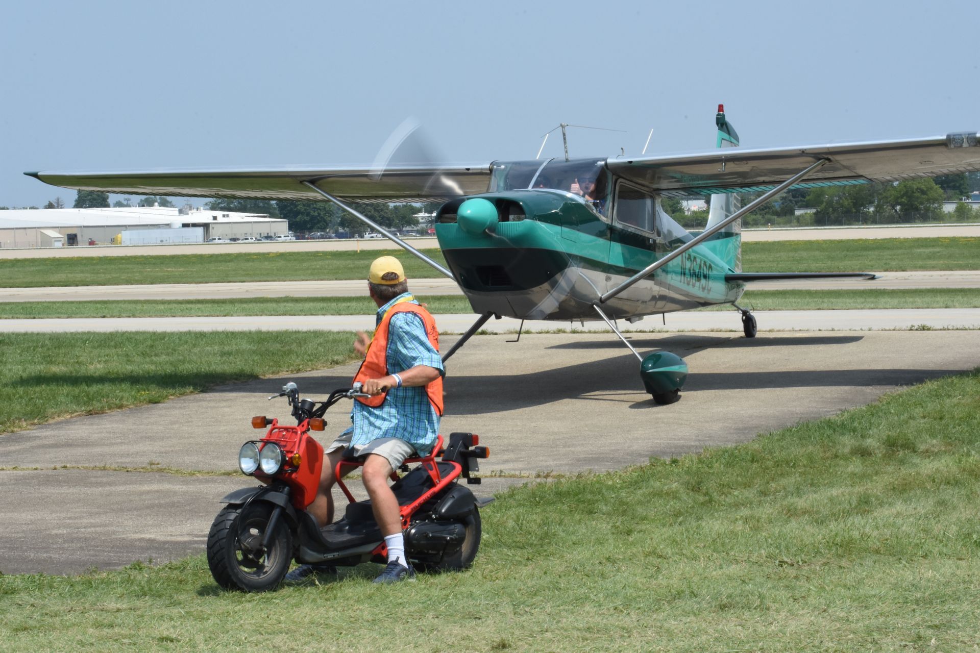 A person wearing an orange vest on a red scooter on grass, facing a small green and white airplane on an airport tarmac.