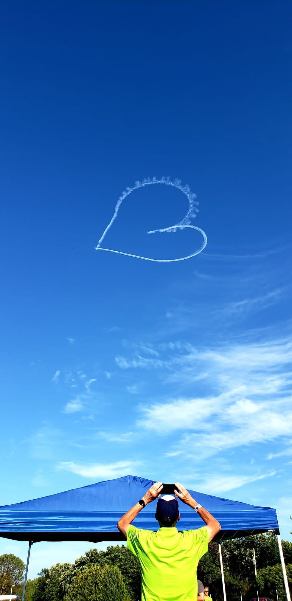 Person in neon shirt making a heart shape overhead under a blue sky with a heart-shaped skywriting trail